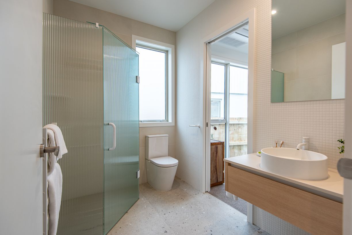 A comprehensive bathroom renovation by Level Plumbing Howick, showcasing a modern back-to-wall toilet suite and a walk-in shower with textured reeded glass doors. The bright, contemporary space includes a light timber vanity with a white vessel basin, sle