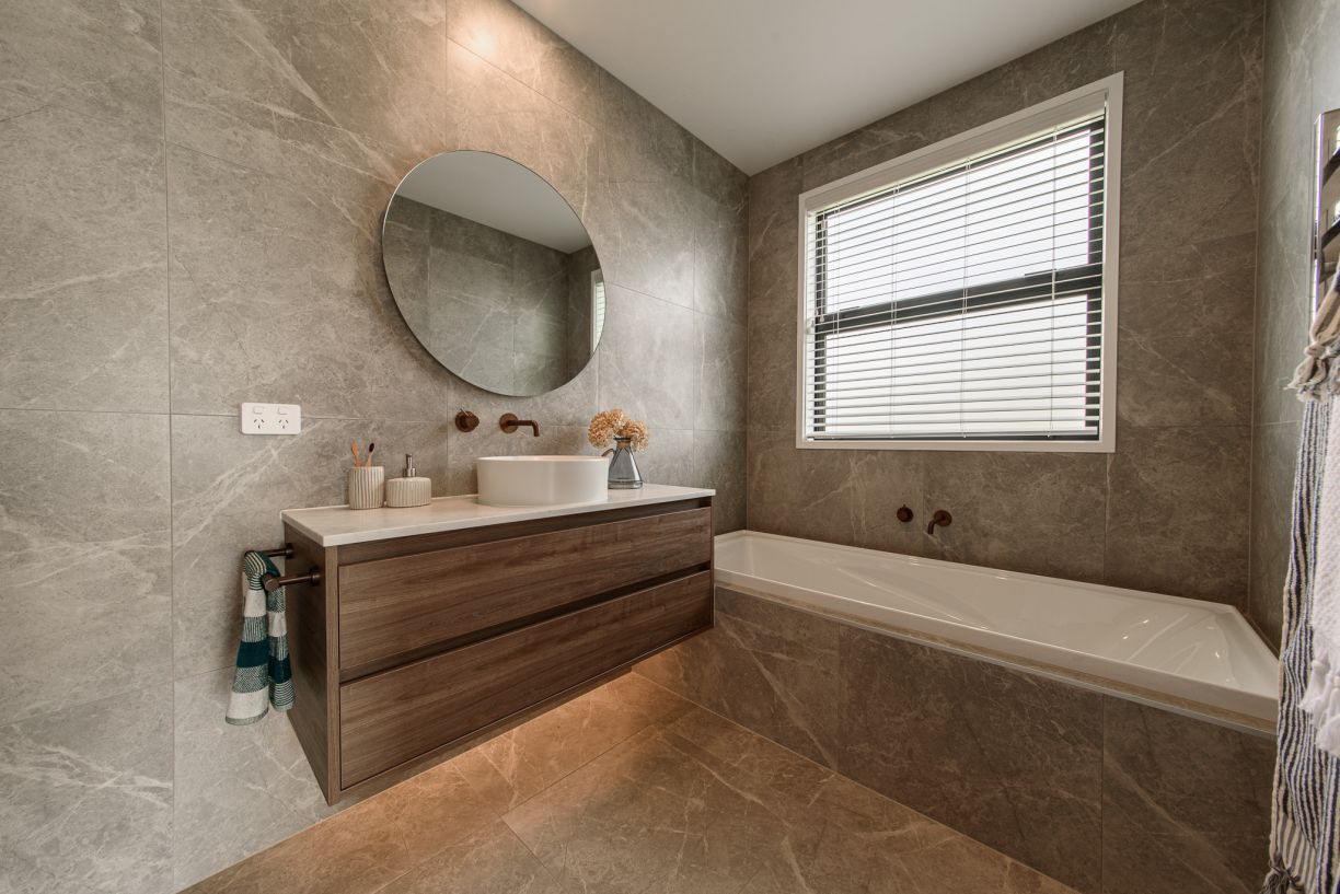 A master bathroom renovation completed by Level Plumbing Howick, showcasing a modern inset bathtub surrounded by grey marble-look tiles and a matching floating timber vanity. Features include a round mirror, a white vessel sink, and wall-mounted copper ta