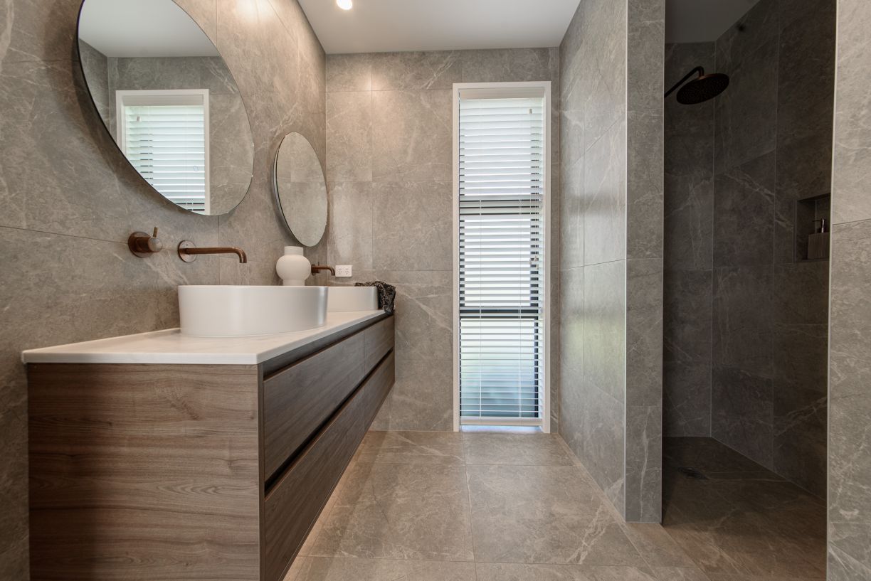 A luxury master bathroom renovation completed by Level Plumbing Howick, showcasing a floating timber double vanity with white vessel sinks and wall-mounted copper tapware. The space features large grey marble-look porcelain wall and floor tiles, round mir