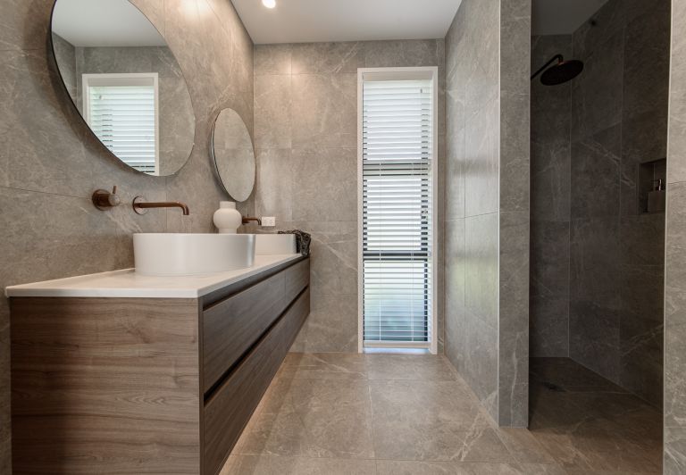 A luxury master bathroom renovation completed by Level Plumbing Howick, showcasing a floating timber double vanity with white vessel sinks and wall-mounted copper tapware. The space features large grey marble-look porcelain wall and floor tiles, round mir
