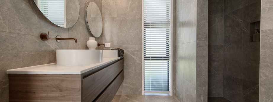 A luxury master bathroom renovation completed by Level Plumbing Howick, showcasing a floating timber double vanity with white vessel sinks and wall-mounted copper tapware. The space features large grey marble-look porcelain wall and floor tiles, round mir