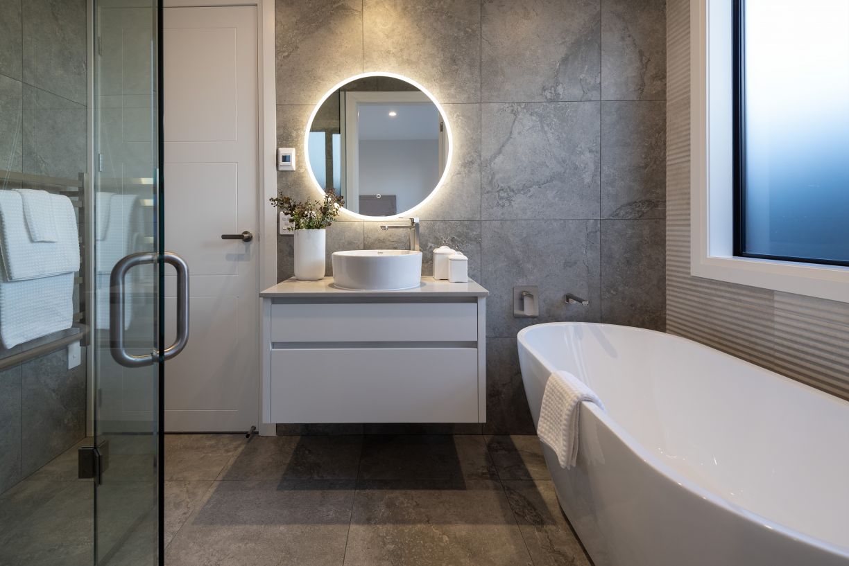 A luxury master bathroom renovation completed by Level Plumbing Howick. The modern space features a white freestanding oval bathtub with wall-mounted chrome tapware, a floating white vanity with a ceramic vessel basin, and a backlit round LED mirror. The 
