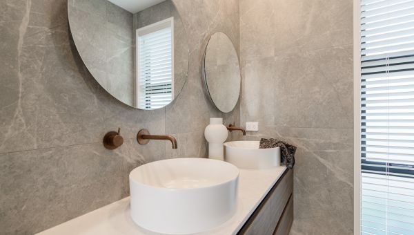 A close-up view of a luxury master bathroom renovation by Level Plumbing Howick, showcasing dual white ceramic vessel sinks on a floating timber double vanity. Features include wall-mounted brushed copper mixer taps, round mirrors, and large-format grey m