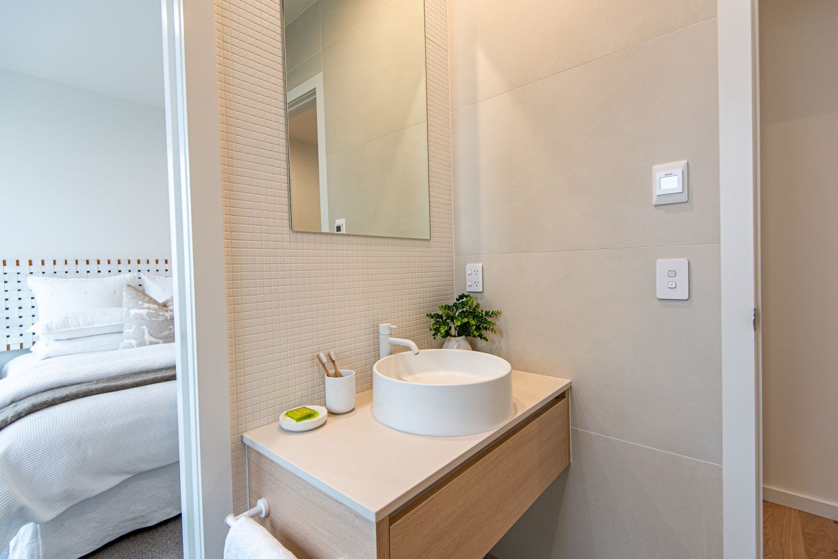 A compact modern ensuite bathroom renovation completed by Level Plumbing Howick. The space features a floating timber vanity with a round white vessel basin and a minimalist white faucet. The wall behind the mirror is lined with small square mosaic tiles,
