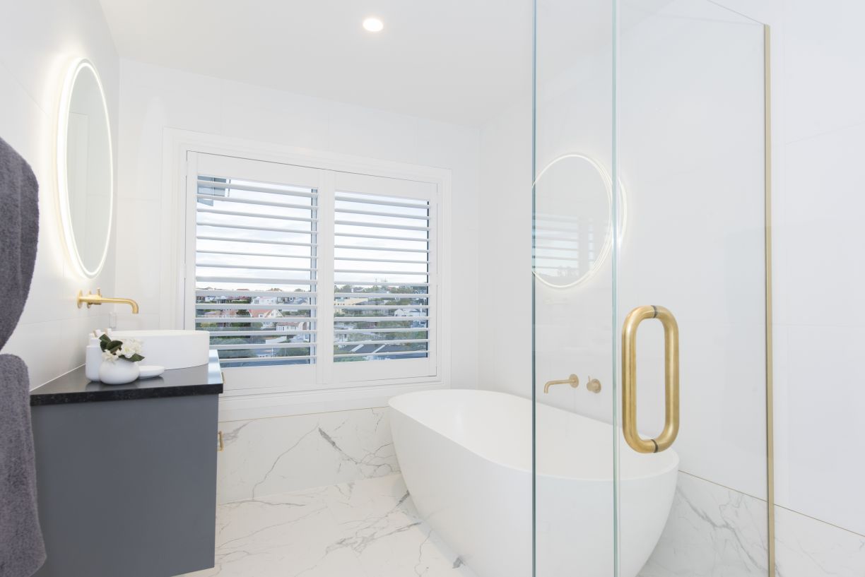 A modern master bathroom renovation by Level Plumbing Howick, showcasing a white freestanding oval bathtub with wall-mounted brass tapware. To the left, a dark grey vanity features a white vessel basin and an illuminated round mirror, all set against marb