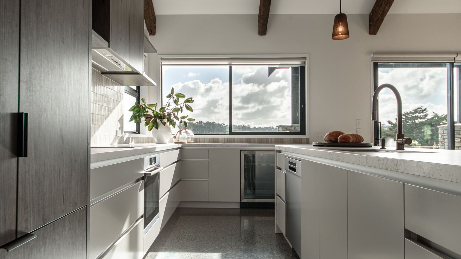 A sleek, contemporary kitchen fit-out by Level Plumbing Howick, featuring a modern matte black gooseneck tap installed on a white engineered stone island bench. The space includes an integrated under-bench dishwasher, premium cabinetry, polished concrete 