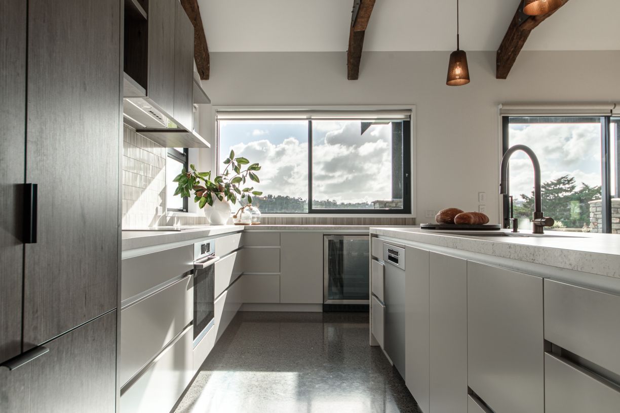 A sleek, contemporary kitchen fit-out by Level Plumbing Howick, featuring a modern matte black gooseneck tap installed on a white engineered stone island bench. The space includes an integrated under-bench dishwasher, premium cabinetry, polished concrete 