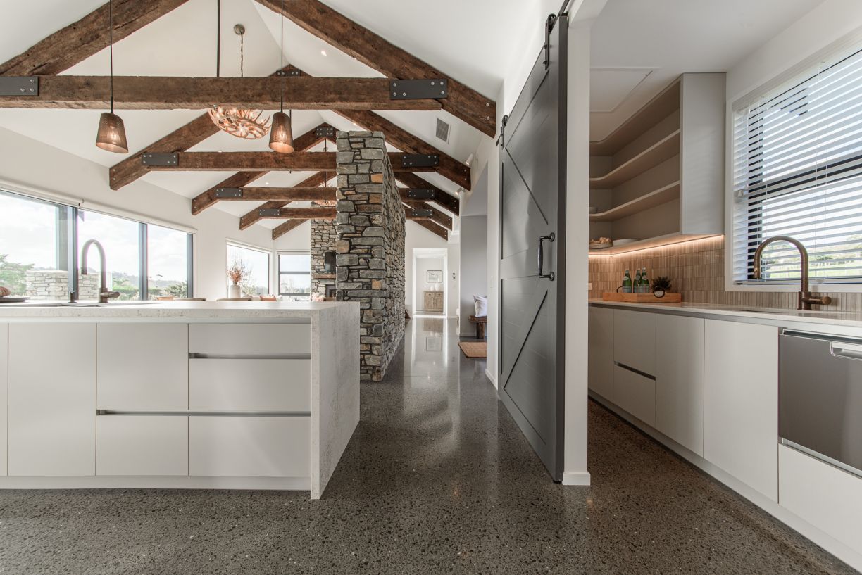 A large open-plan luxury kitchen and adjacent scullery installed by Level Plumbing Howick. The modern space features polished concrete floors, exposed timber ceiling beams, white cabinetry, a stone kitchen island with a gooseneck tap, and a separate butle