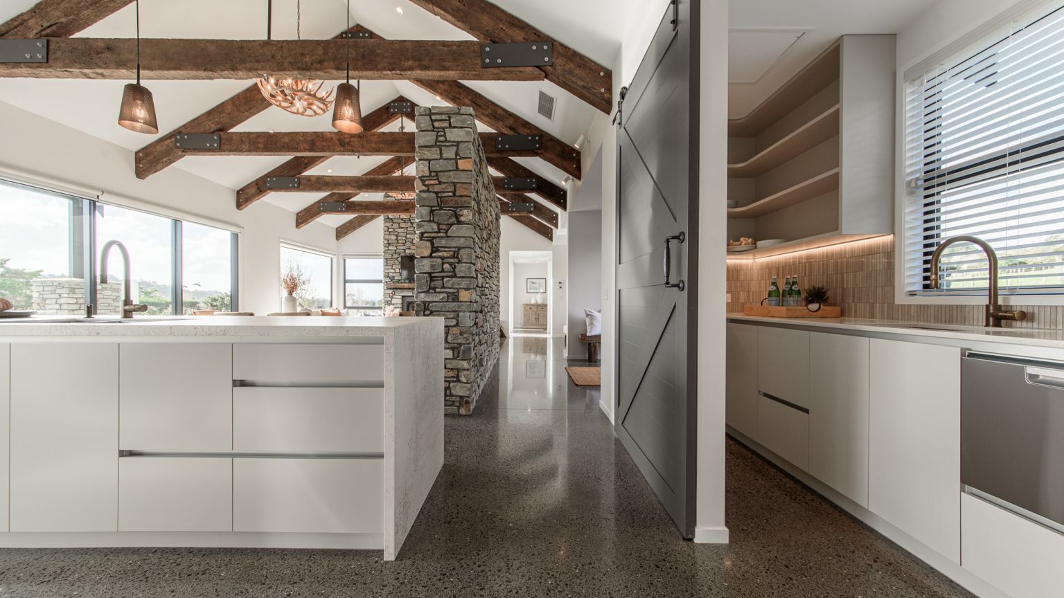 A large open-plan luxury kitchen and adjacent scullery installed by Level Plumbing Howick. The modern space features polished concrete floors, exposed timber ceiling beams, white cabinetry, a stone kitchen island with a gooseneck tap, and a separate butle