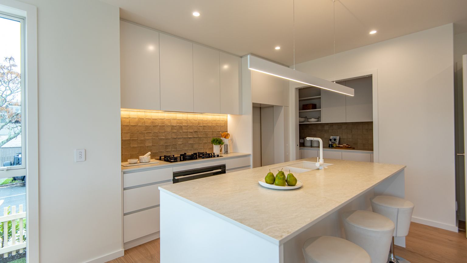 A contemporary kitchen and scullery installation completed by Level Plumbing Howick. The open-plan space features a white kitchen island with a beige engineered stone countertop, an undermount double sink, and a modern white mixer tap. The background show