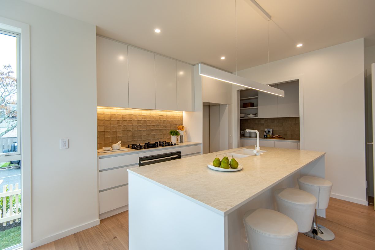 A contemporary kitchen and scullery installation completed by Level Plumbing Howick. The open-plan space features a white kitchen island with a beige engineered stone countertop, an undermount double sink, and a modern white mixer tap. The background show