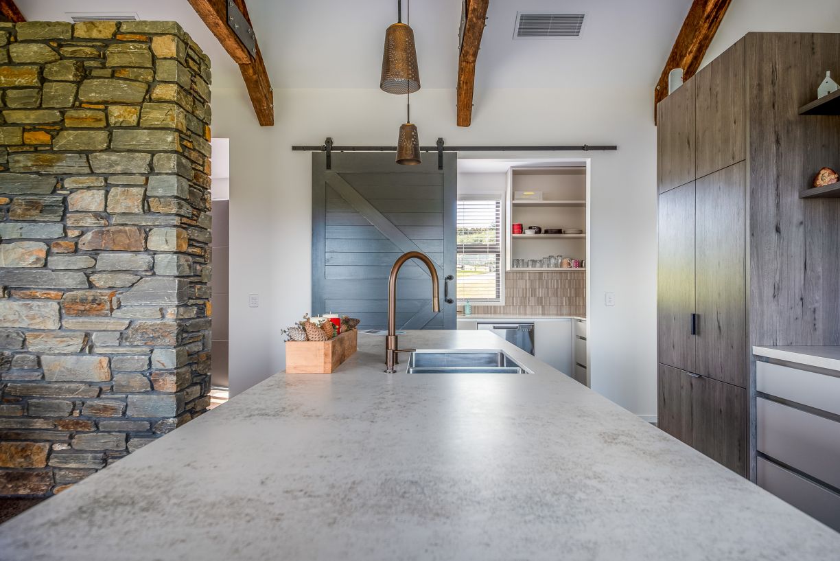 A close-up view of a luxury kitchen island installation by Level Plumbing Howick, featuring an undermount stainless steel sink with a premium copper gooseneck faucet. The background shows a stone accent pillar, wood beams, and a glimpse into a butler's pa