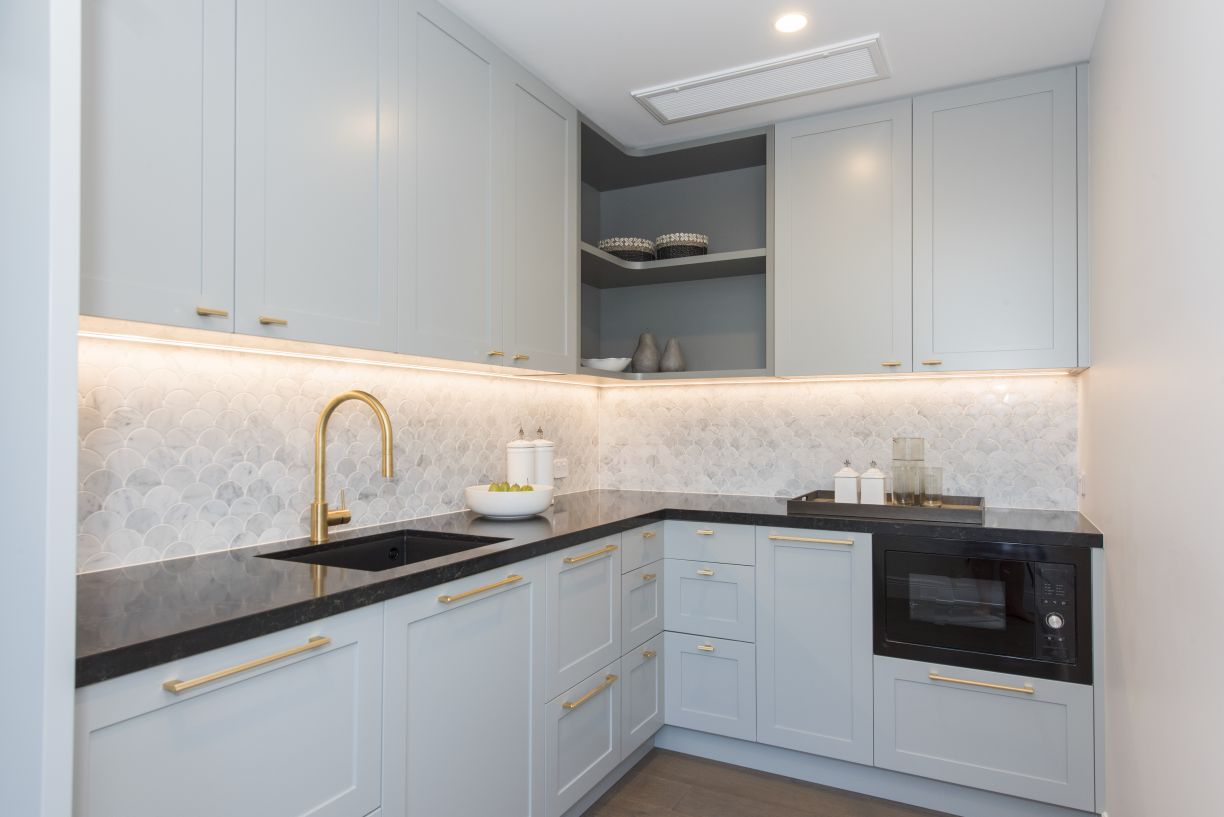 A luxury kitchen fit-out by Level Plumbing Howick, featuring a modern drop-in black sink paired with a premium brushed brass gooseneck faucet. The space is styled with duck-egg blue shaker cabinets, dark stone benchtops, brass handles, and a white fish-sc