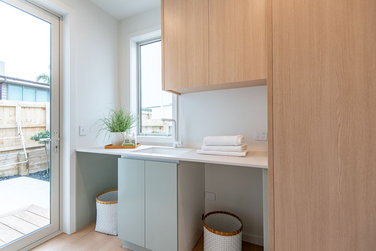 A modern laundry room fit-out completed by Level Plumbing Howick, featuring a drop-in white laundry tub sink and a minimalist white mixer tap. The space includes a white countertop with openings for an under-bench washing machine and dryer, soft sage gree
