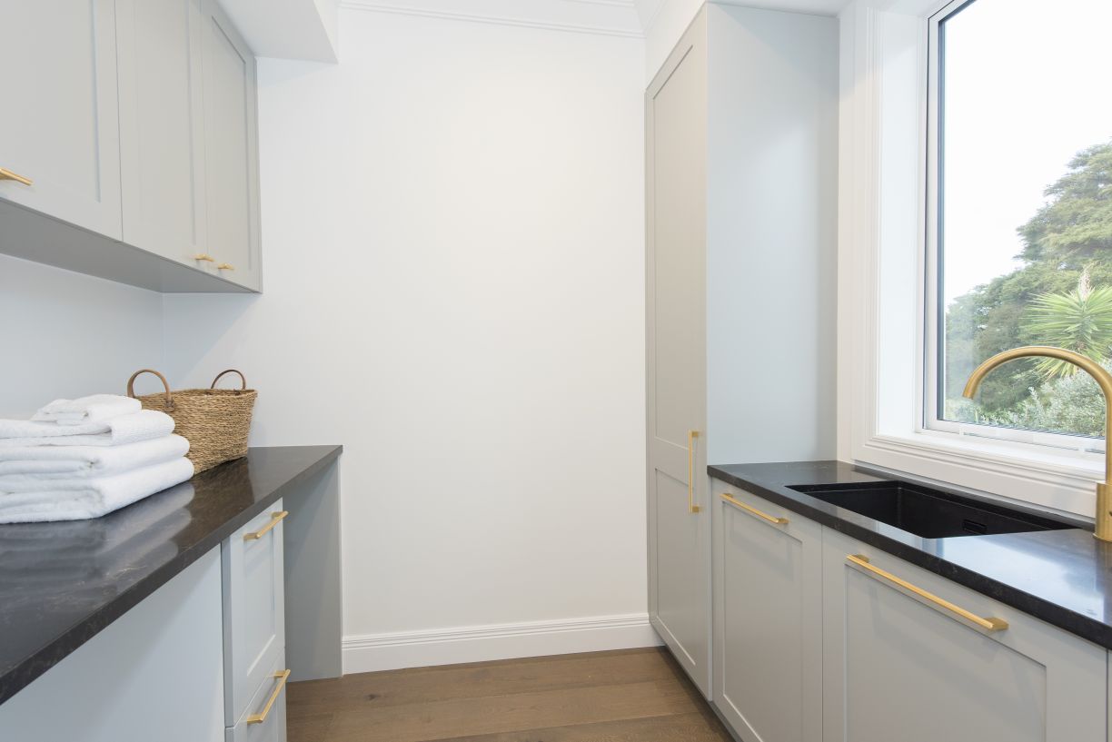 A sleek laundry room renovation completed by Level Plumbing Howick, showcasing an undermount black laundry tub with a brushed brass mixer tap positioned under a large window. The room features light grey cabinetry with brass hardware, dark countertops, an