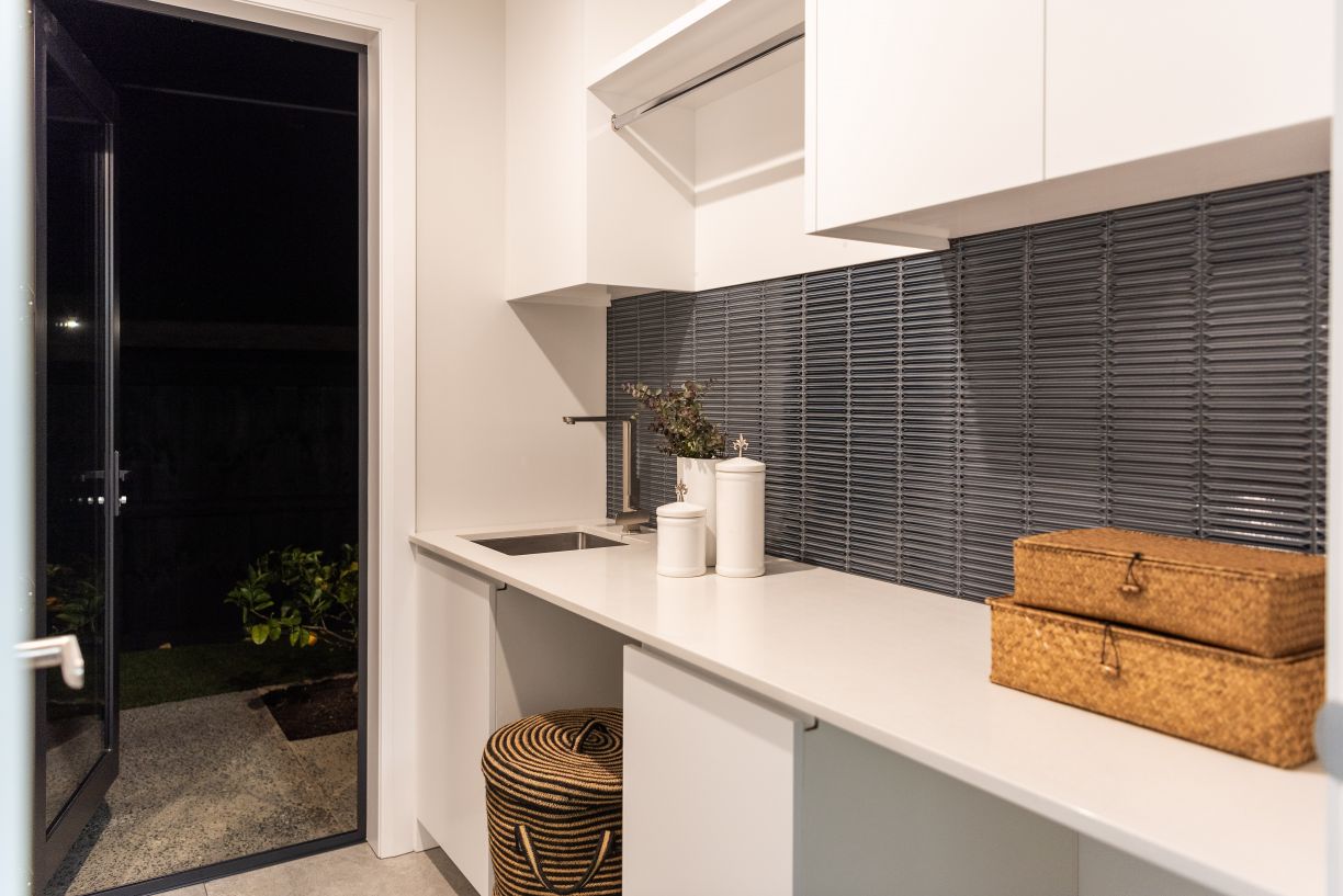 A modern laundry room fit-out completed by Level Plumbing Howick, showcasing an undermount stainless steel utility sink and a sleek chrome tap. The clean workspace features white stone countertops, white upper and lower cabinetry, a dark grey kit-kat mosa