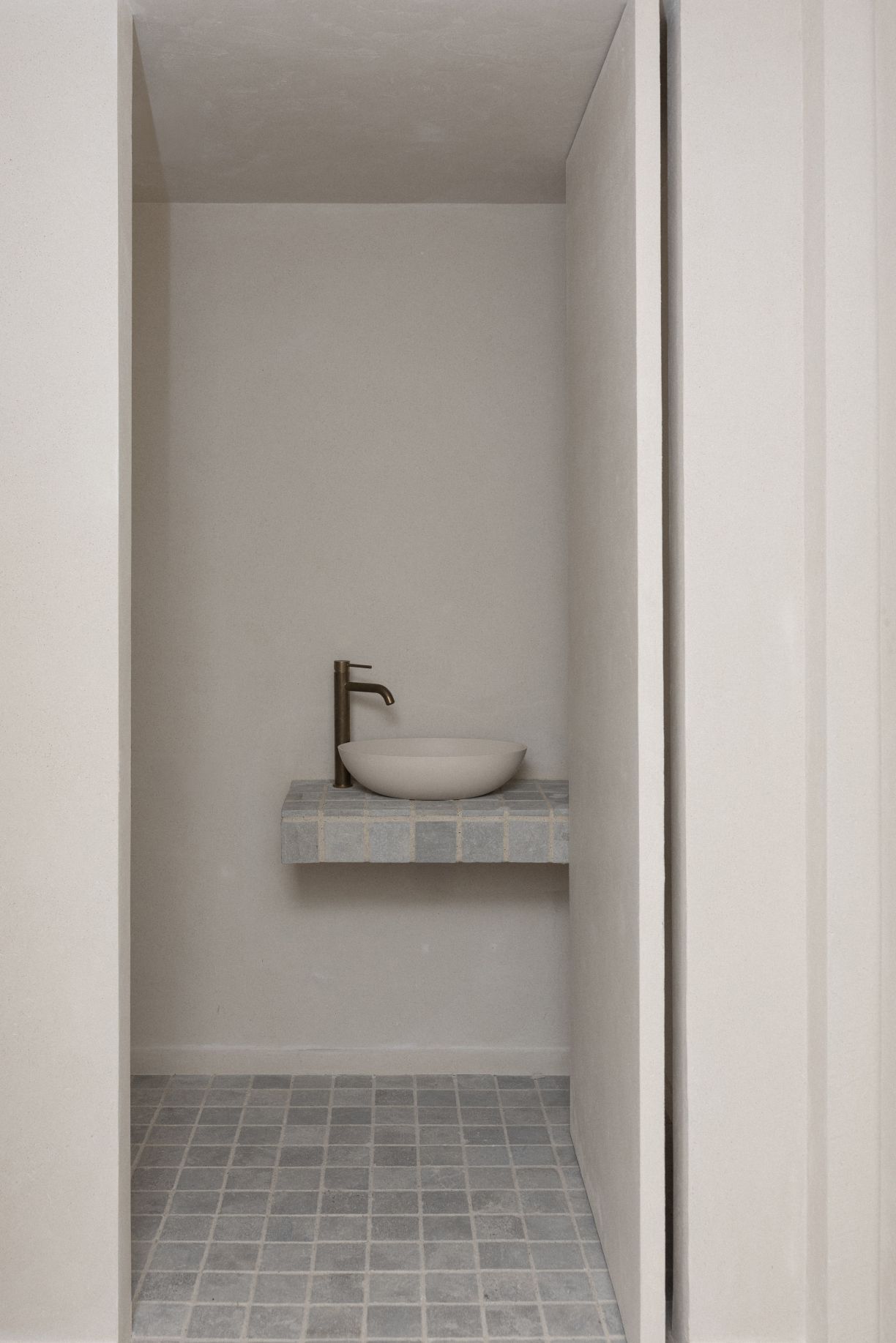 A minimalist modern bathroom renovation in Howick, Auckland, featuring a custom tiled floating vanity counter, light stone vessel sink, and an elegant brass tap against textured plaster walls.