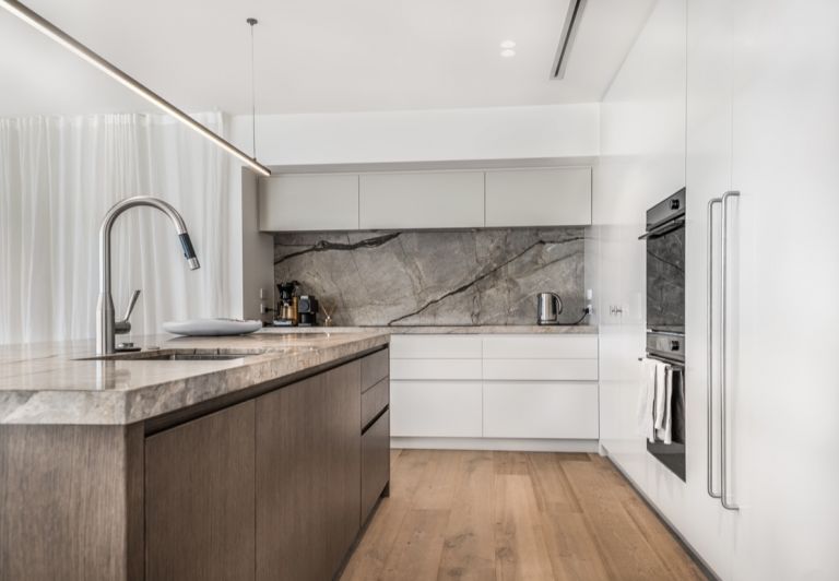 A luxury modern kitchen renovation in Howick, Auckland, showcasing a high-end gooseneck pull-out mixer tap installed on a marble island bench. The background features sleek white and timber cabinetry with a matching stone slab splashback.
