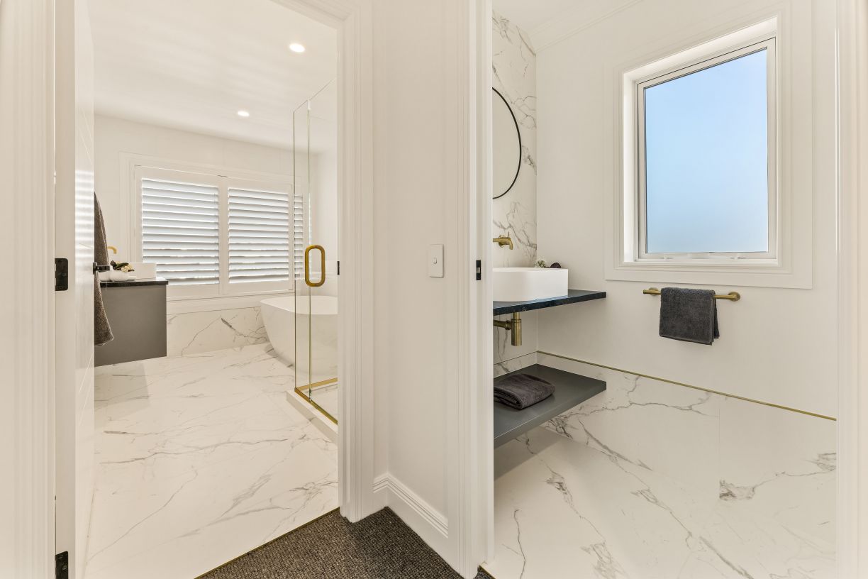 A high-end master bathroom and adjacent powder room renovation completed by Level Plumbing Howick. The modern spaces feature matching white marble-look tile floors and wall accents, paired with premium brass tapware and fittings. The powder room shows a f