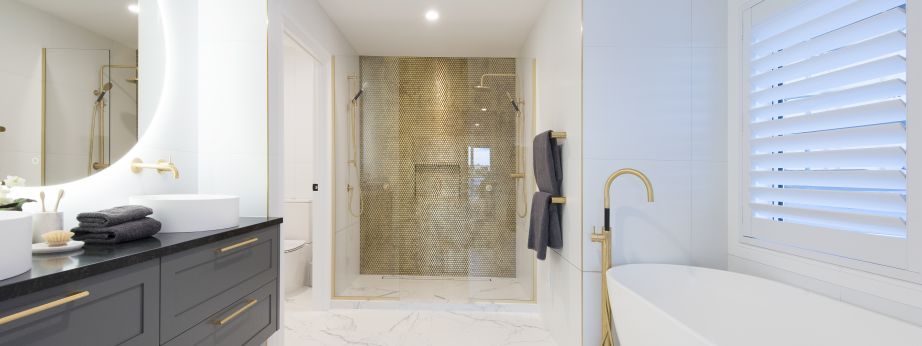 A full view of a luxury bathroom installation completed by Level Plumbing Howick. The space features a white freestanding bath with a floor-mounted brass filler tap, a large dark grey double vanity with dual vessel sinks, and a walk-in shower enclosure hi