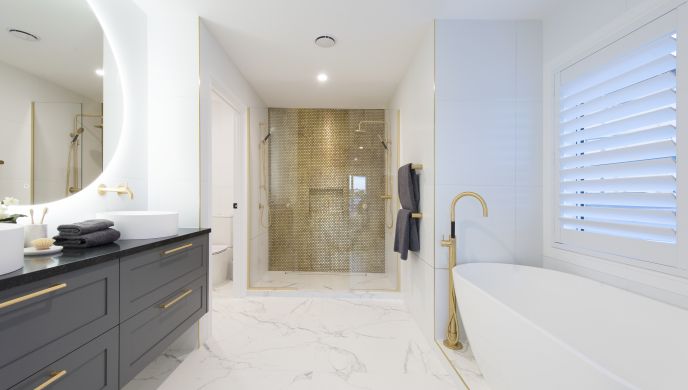 A full view of a luxury bathroom installation completed by Level Plumbing Howick. The space features a white freestanding bath with a floor-mounted brass filler tap, a large dark grey double vanity with dual vessel sinks, and a walk-in shower enclosure hi