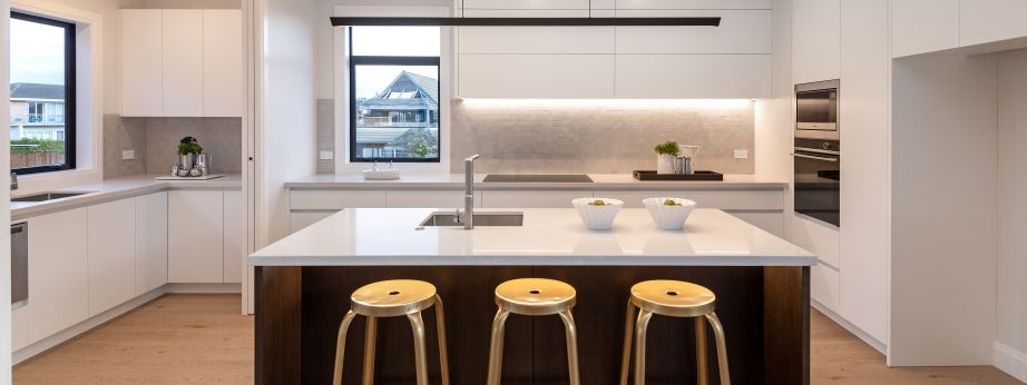 A straight-on view of a luxury kitchen and scullery renovation completed by Level Plumbing Howick. The open-plan layout showcases a dark timber island bench with a white stone countertop, an undermount square stainless steel sink, a modern chrome mixer ta