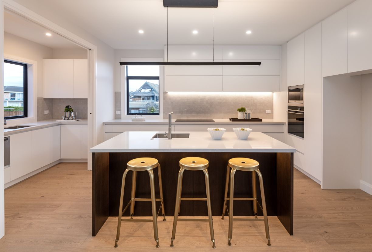 A straight-on view of a luxury kitchen and scullery renovation completed by Level Plumbing Howick. The open-plan layout showcases a dark timber island bench with a white stone countertop, an undermount square stainless steel sink, a modern chrome mixer ta