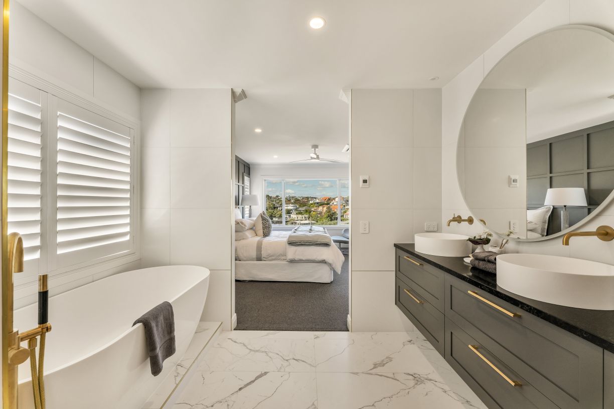 A luxury master ensuite bathroom renovation completed by Level Plumbing Howick. The open layout features a dark grey double vanity with dual white vessel basins and wall-mounted brass tapware beneath a large round mirror. To the left, a white freestanding