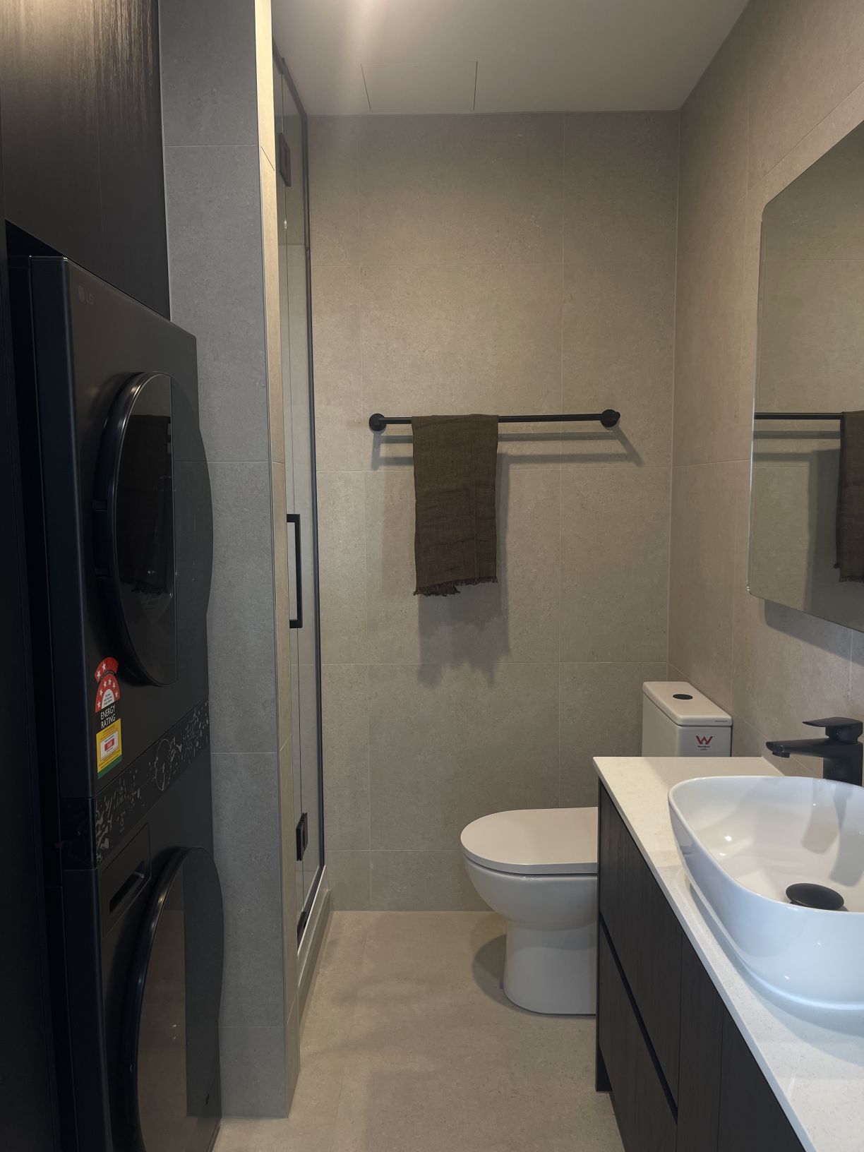 Modern bathroom and laundry space featuring stacked front-load laundry appliances, back-to-wall toilet, and floating vanity with black tapware installed by Level Plumbing Howick.