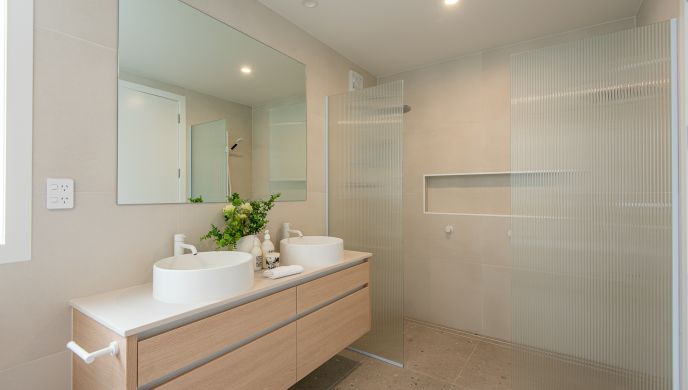 A contemporary master bathroom renovation completed by Level Plumbing Howick, featuring a light oak double vanity with dual white vessel sinks and modern white tapware. The space is styled with beige wall tiles, terrazzo-style floor tiles, a large rectang
