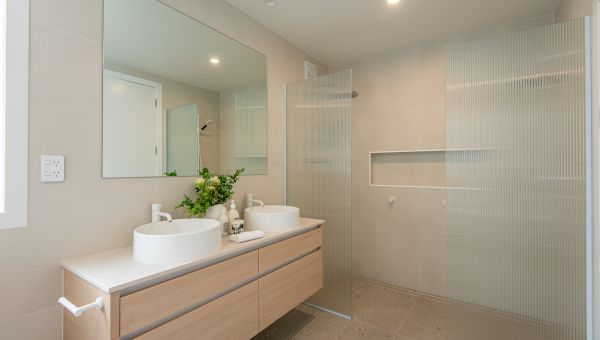 A contemporary master bathroom renovation completed by Level Plumbing Howick, featuring a light oak double vanity with dual white vessel sinks and modern white tapware. The space is styled with beige wall tiles, terrazzo-style floor tiles, a large rectang