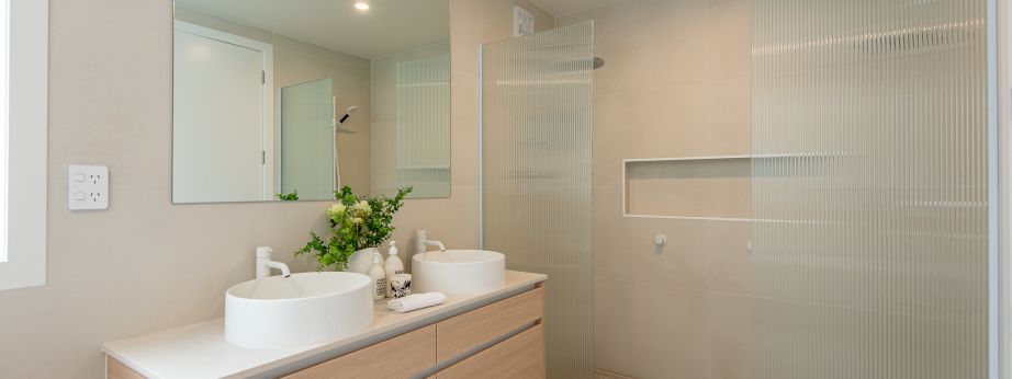 A contemporary master bathroom renovation completed by Level Plumbing Howick, featuring a light oak double vanity with dual white vessel sinks and modern white tapware. The space is styled with beige wall tiles, terrazzo-style floor tiles, a large rectang