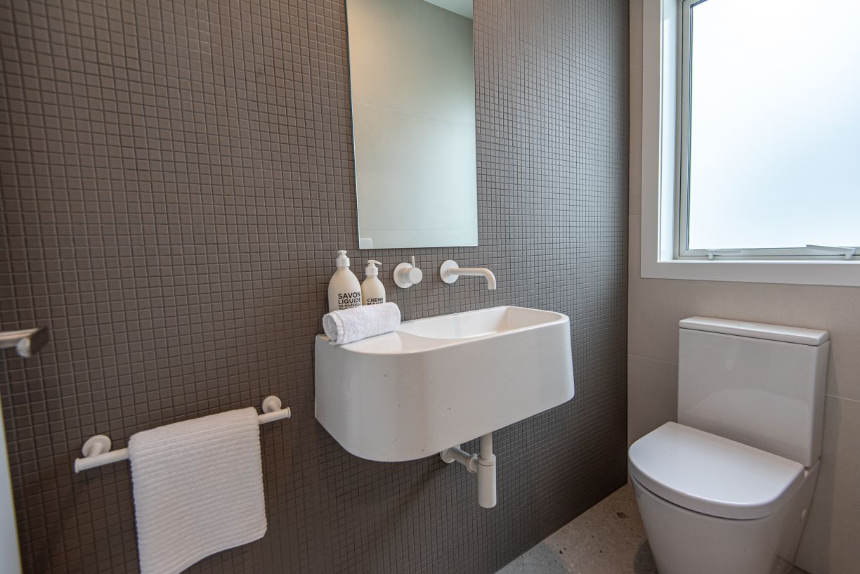A contemporary guest powder room renovation by Level Plumbing Howick, showcasing a wall-hung white stone basin with wall-mounted white tapware and a matching white bottle trap. The space features a dark grey mosaic tiled feature wall, a modern back-to-wal