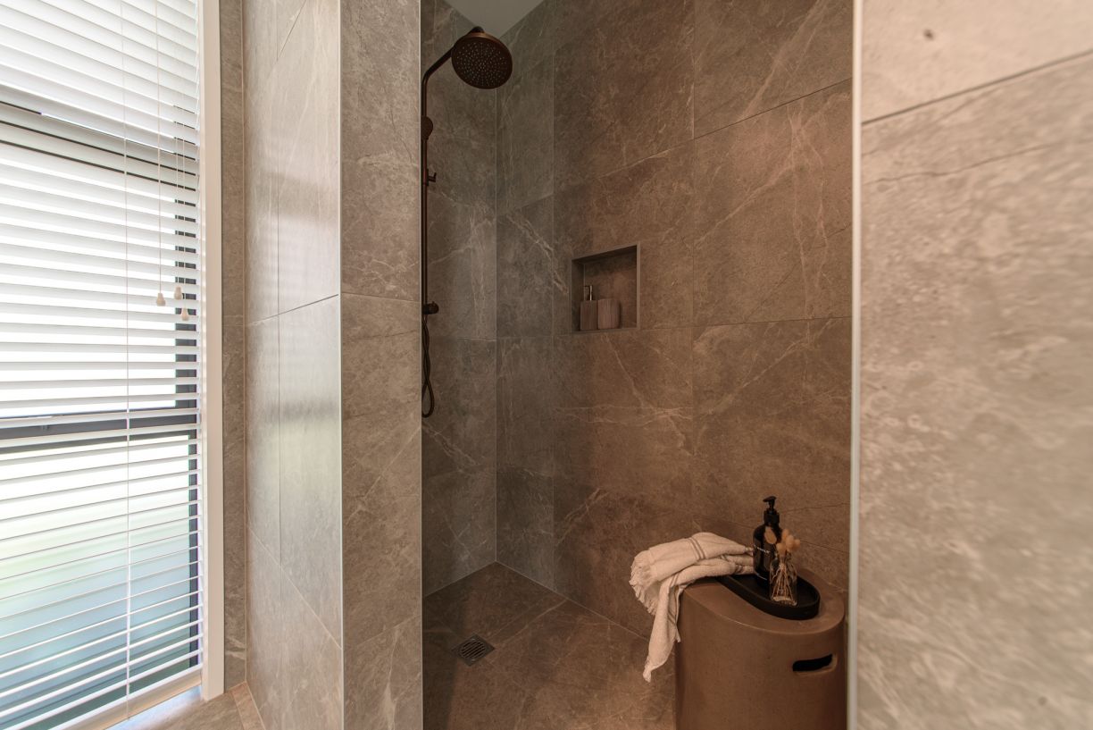A modern walk-in tiled shower enclosure installed by Level Plumbing Howick, featuring an overhead copper rainfall shower rose and matching mixer valve. The space is fully lined with grey marble-look porcelain tiles and includes a recessed wall niche next 